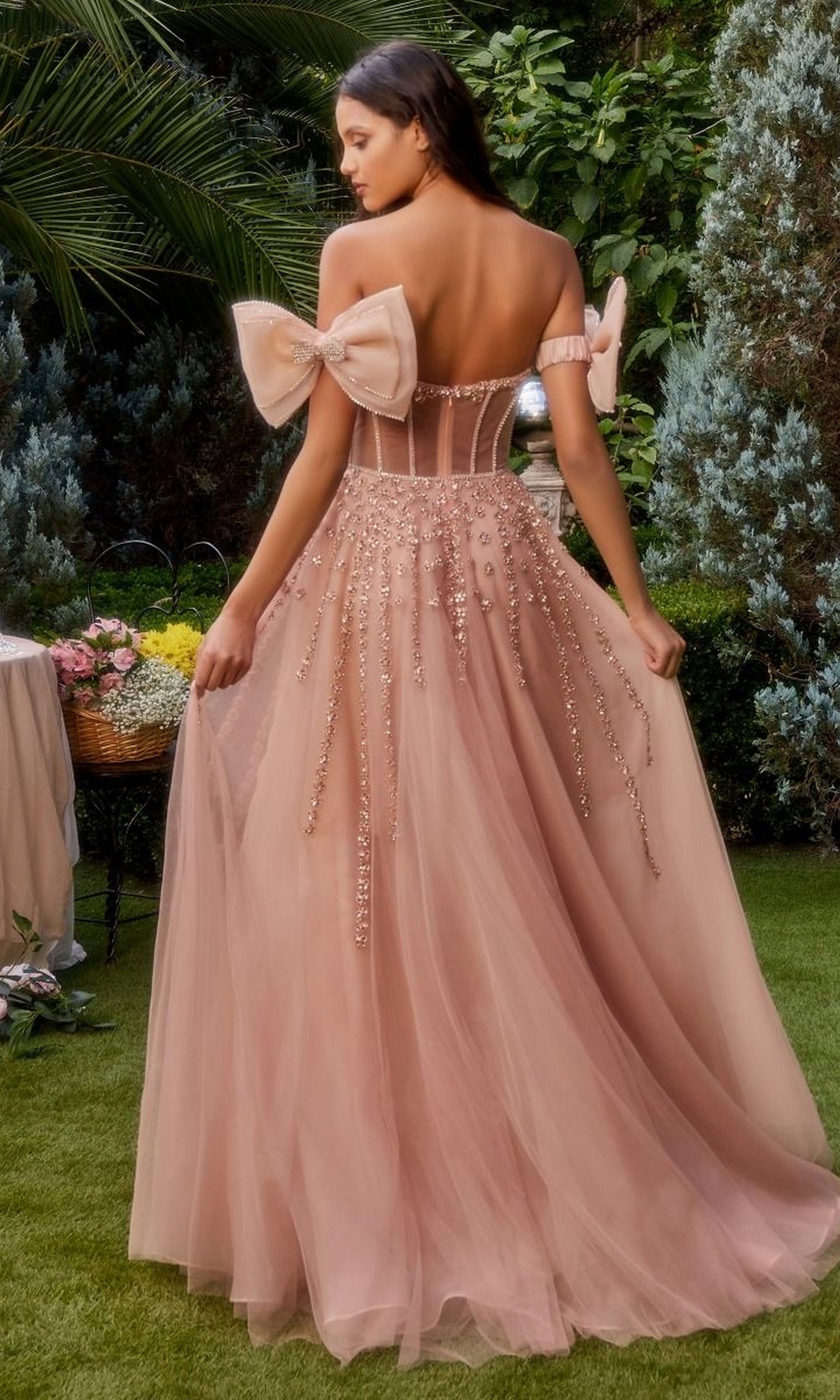 Outdoors, a woman wears the Andrea and Leo A1338 strapless long prom dress in blush pink, featuring beaded details, a tulle skirt, and large bows on the sleeves.